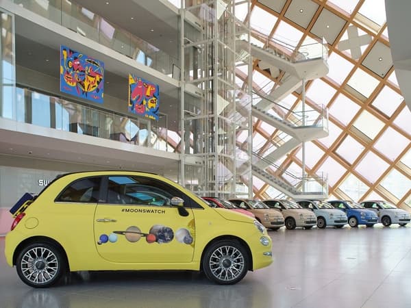 Sebuah FIAT 500 berwarna kuning dengan tulisan #MOONSWATCH dan grafis planet di pintunya, terparkir di depan deretan mobil FIAT 500 berwarna-warni