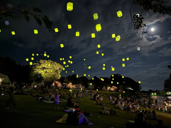 Lanterns floating in the night sky above people seated on the lawn at Resol no Mori