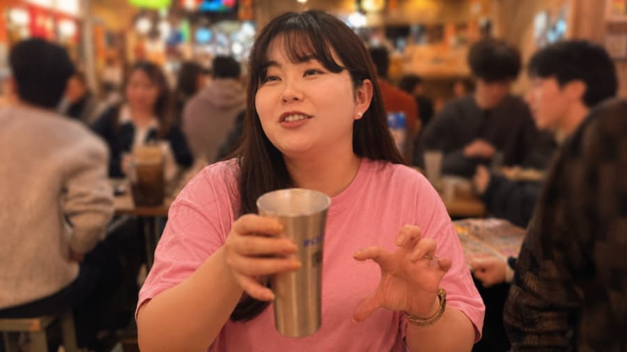 Sayaka talking with a metal tumbler in hand at a busy izakaya full of people