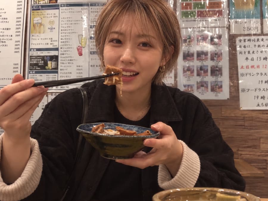 Rina smiling and holding up a bite of simmered food on chopsticks in front of handwritten izakaya menus
