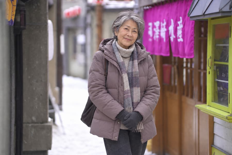 Eri in a down jacket and plaid scarf smiling in a snowy alley lined with shop curtains