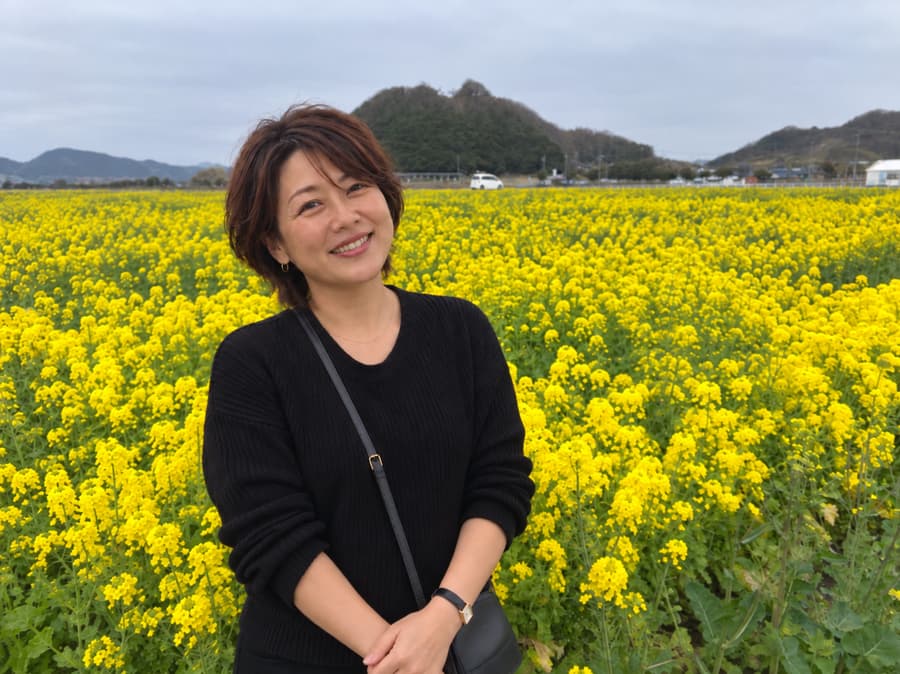 Maika smiling in a black sweater in front of a field of yellow rapeseed blossoms