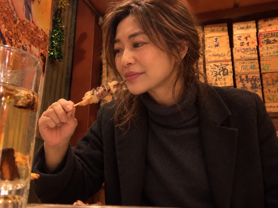 Kana smiling as she tastes a skewer at a cozy bar lined with handwritten wooden plaques