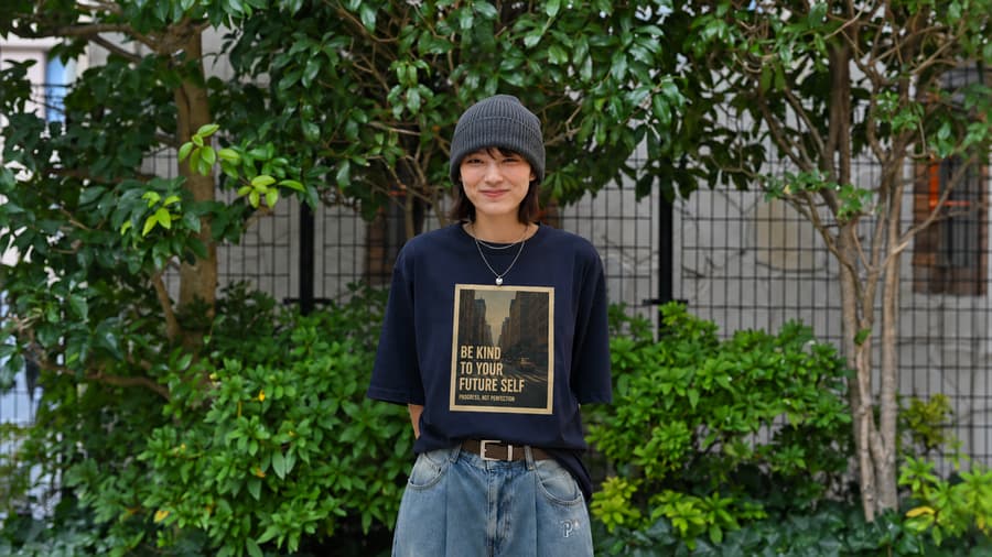 Kanna in a beanie and printed tee standing in front of a hedge