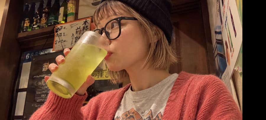 Saki in glasses and a knit beanie sipping a highball at a cozy izakaya with handwritten menus