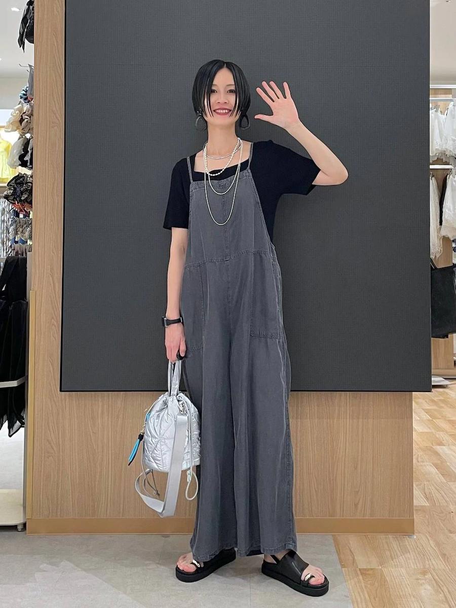 Rina waving in grey denim dungarees, standing in a clothing shop