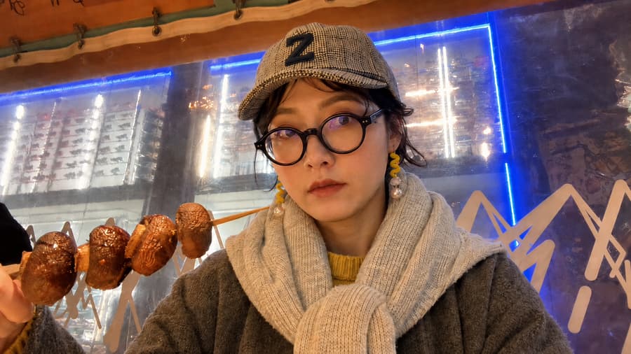 Honoka in a check cap and round glasses beside a skewer of grilled food at a night stall