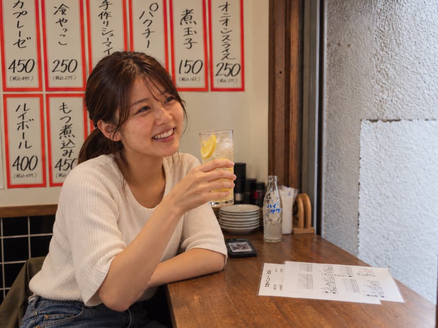 Mari smiling with a lemon sour at a wooden counter beneath handwritten menu cards