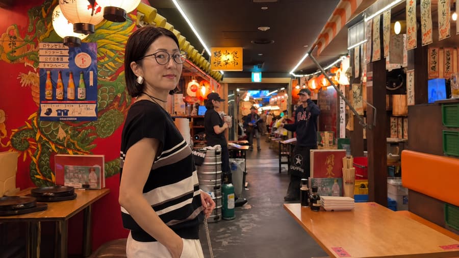 Sae standing in a lantern-lit alley lined with izakaya stalls