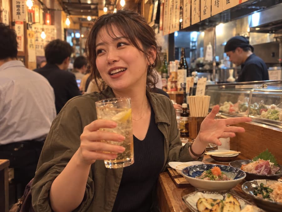 Aya laughing with a highball in hand at a busy izakaya counter lined with small plates