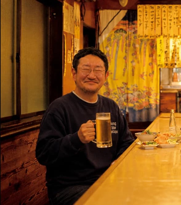 Yoshito smiling toward the camera with a beer mug at a cozy izakaya counter