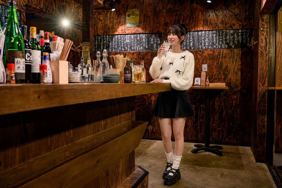 Ayaka standing at a wooden izakaya counter with a glass in hand and sake bottles lined up behind