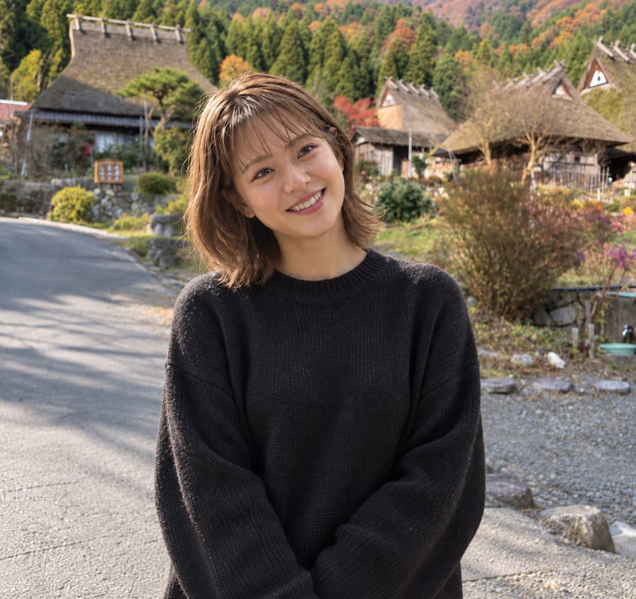 Satoko smiling in a black sweater in front of thatched-roof farmhouses and autumn hills