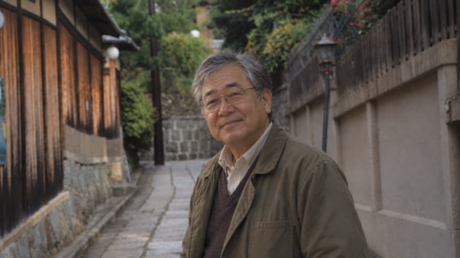 Koji in glasses and an olive jacket smiling on a quiet stone-paved lane lined with old wooden houses