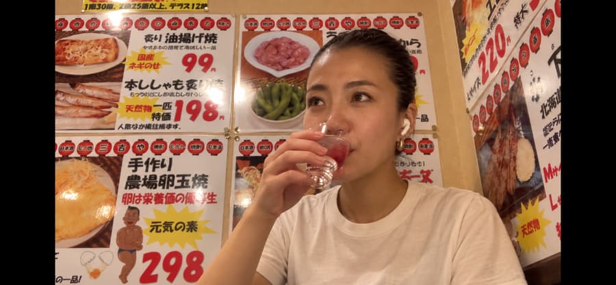 Miyu sipping a drink at an izakaya table in front of hand-written menu posters