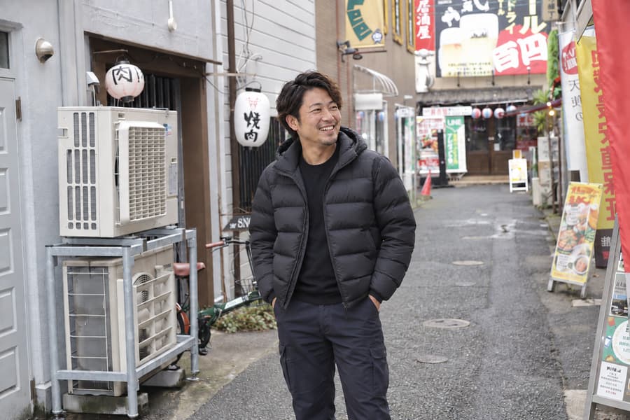 Shohei laughing in a black puffer jacket on a narrow street lined with izakaya lanterns and signs