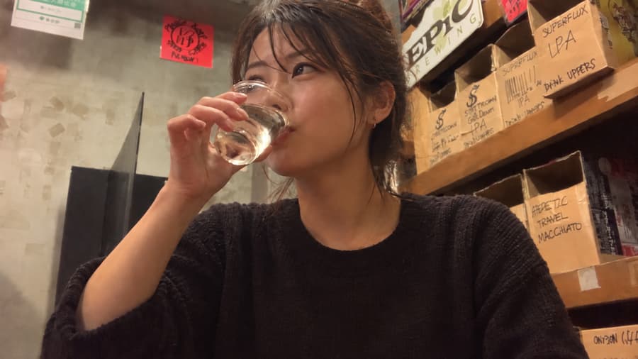 Ema sipping a glass of sake at a bar stacked with handwritten craft beer boxes