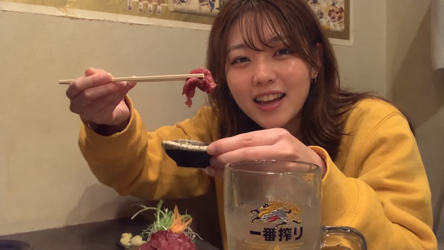Nana smiling and holding up a piece of sashimi on chopsticks behind a large mug at a cozy izakaya table
