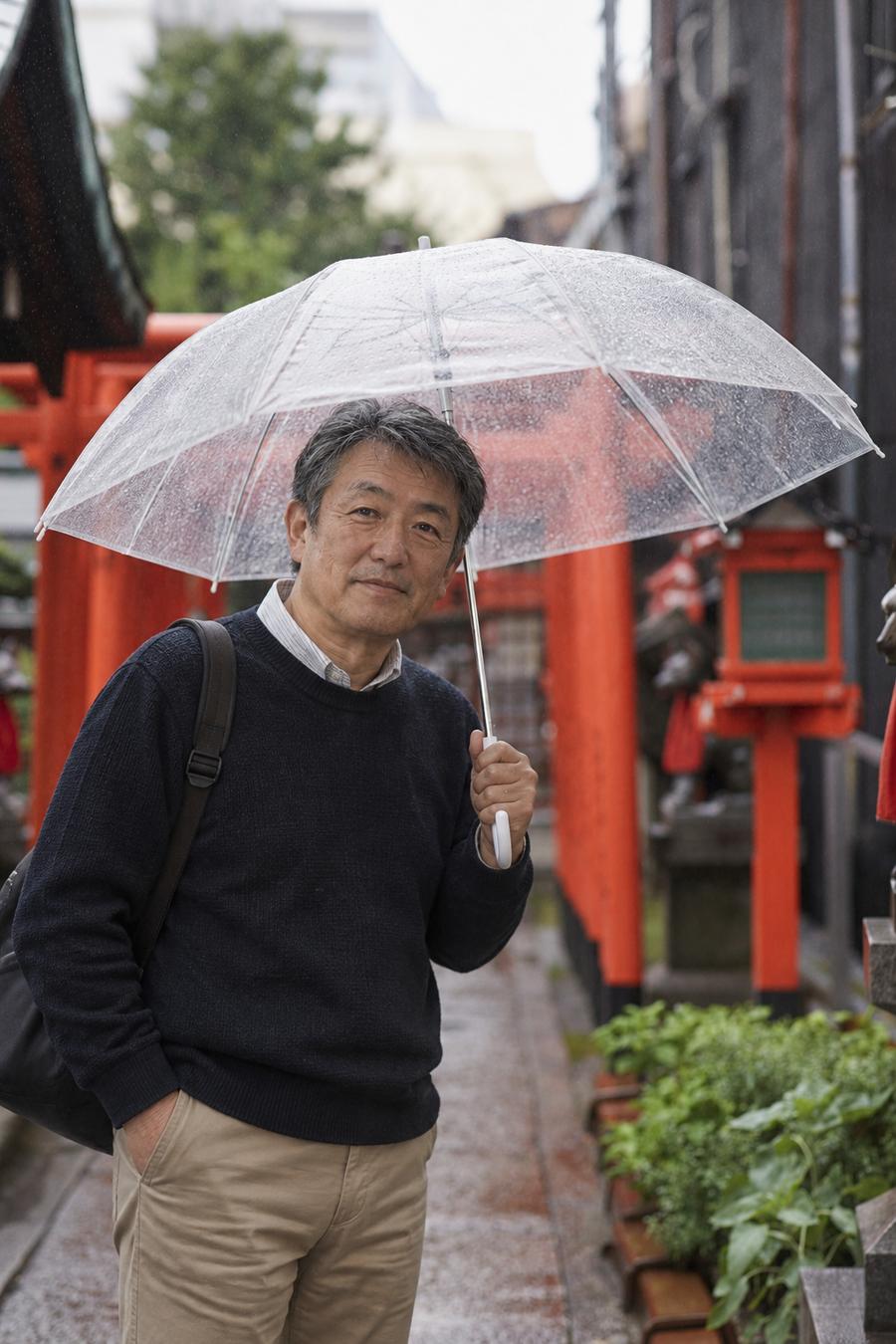 Hiro holding a clear umbrella beside a row of red torii gates on a rainy shrine path