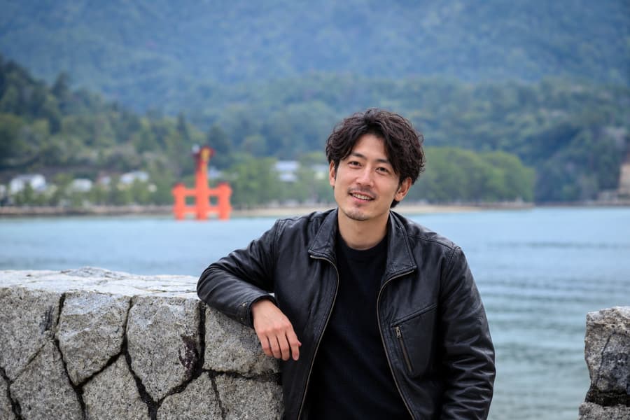 Kai in a black leather jacket leaning on a stone wall by the sea, with a red torii gate in the distance