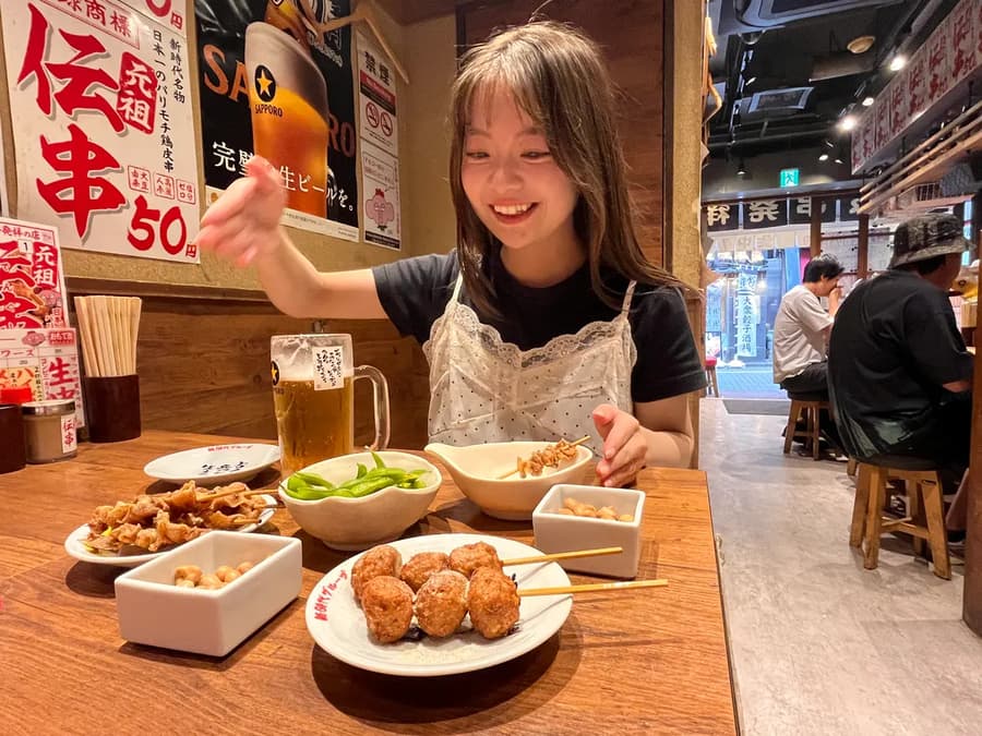 Rena smiling over a table of yakitori and a beer at a lively izakaya