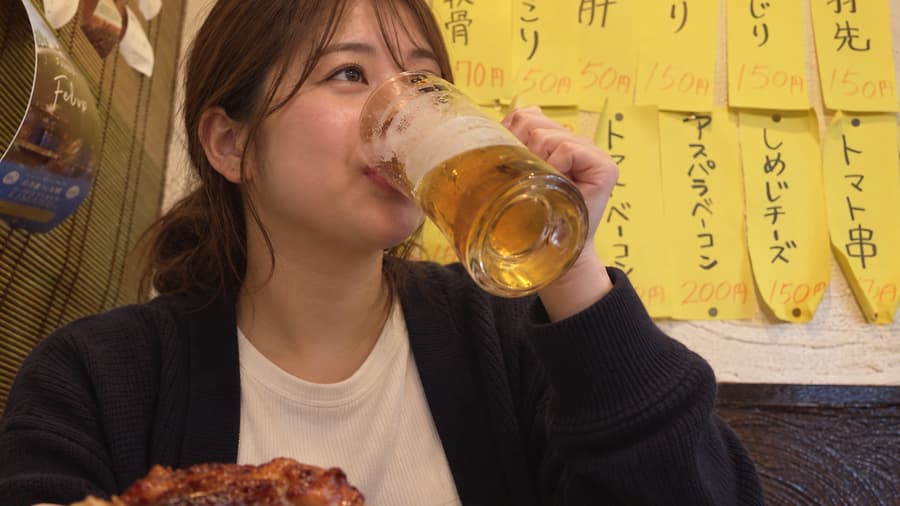Yuka drinking a mug of beer in front of handwritten yakitori menus at an izakaya