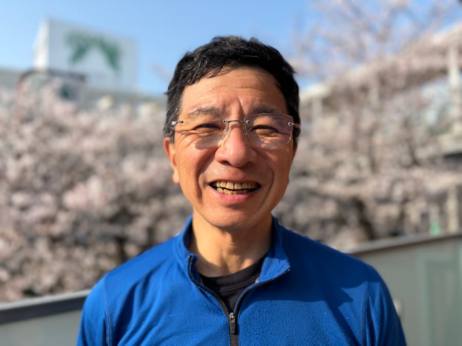 Takeo smiling in a blue fleece with cherry blossoms behind him