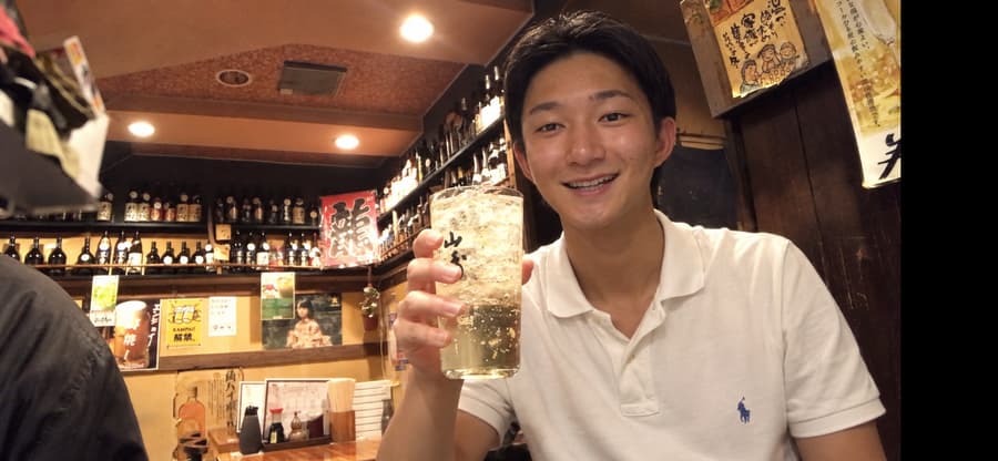 Kanta smiling and raising a highball at a cozy izakaya lined with bottles