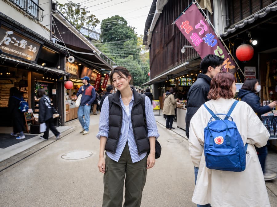 Yui smiling in a striped shirt and black down vest on a lively shopping street lined with red lanterns