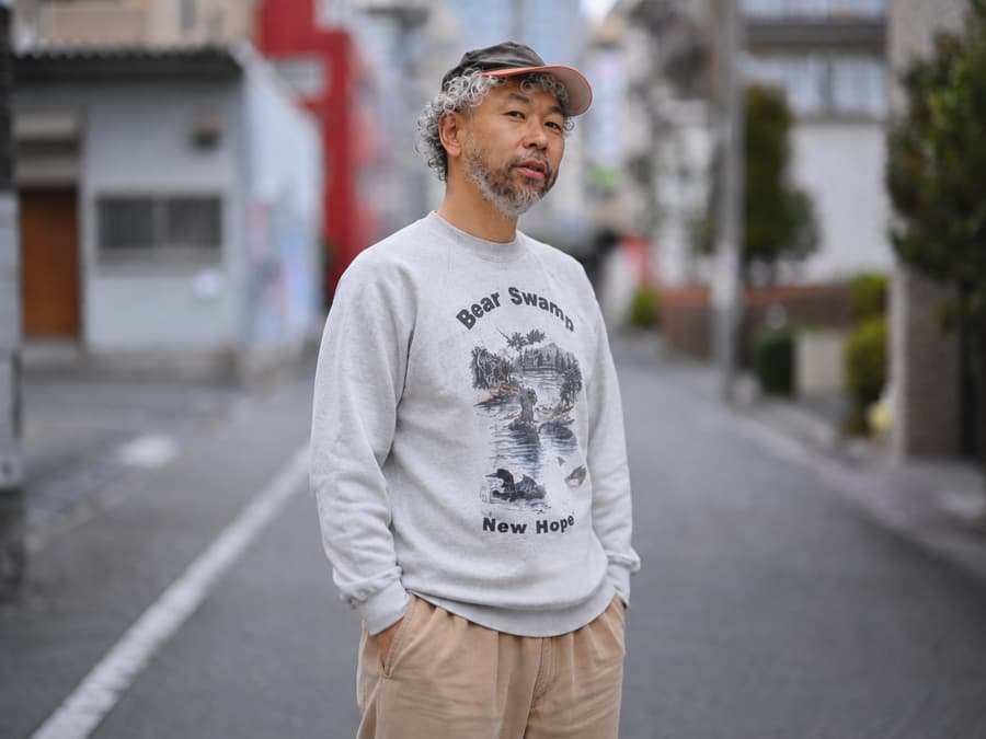 Jin standing on a quiet Tokyo backstreet in a printed sweatshirt and cap, grey hair and beard