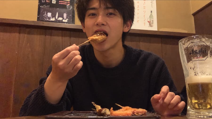 Toma biting into a kushikatsu skewer beside a mug of draft beer at an izakaya