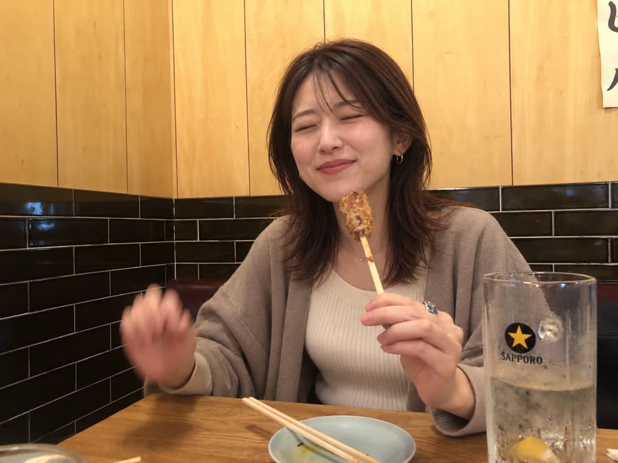 Nozomi smiling with her eyes closed as she holds up a grilled skewer beside a highball at an izakaya table
