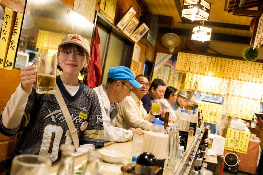 Mika raising a beer mug at a lively old izakaya counter full of regulars