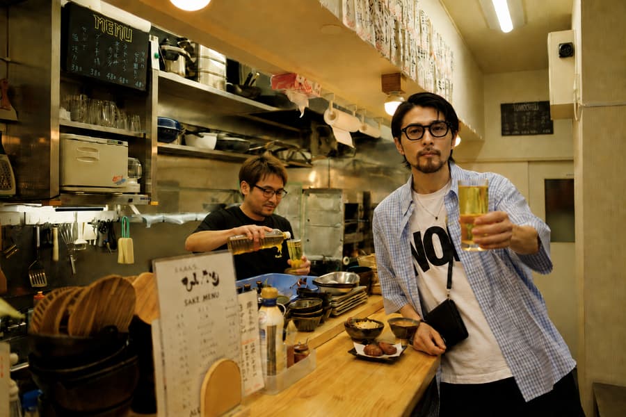 Gen raising a glass of beer at a small izakaya counter while the owner pours behind it