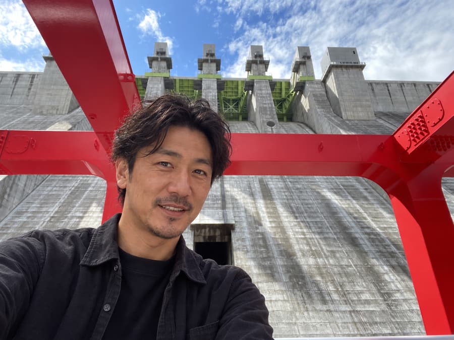 Kenta smiling for a selfie beneath red steel beams in front of a towering concrete dam