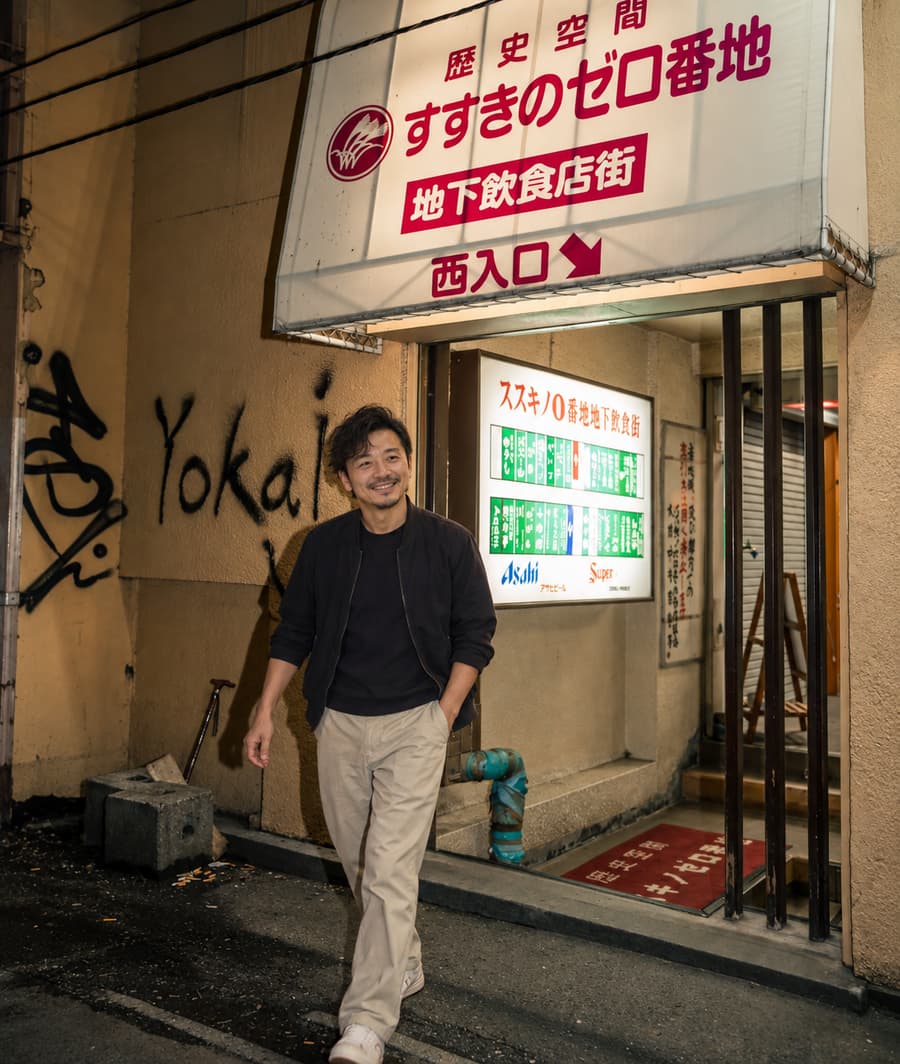 Takumi smiling as he walks past the entrance to an underground dining arcade in Susukino at night