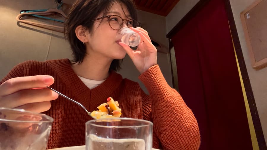 Yuki in glasses and an orange sweater sipping from a small glass, a spoonful of dessert in her other hand