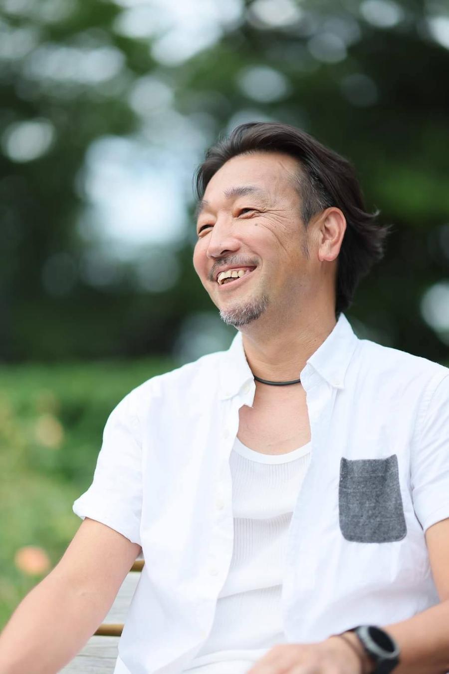 DUKE laughing in a white shirt on a park bench in summer light
