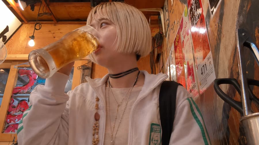 Aoi drinking a tall glass of beer at a poster-covered izakaya counter