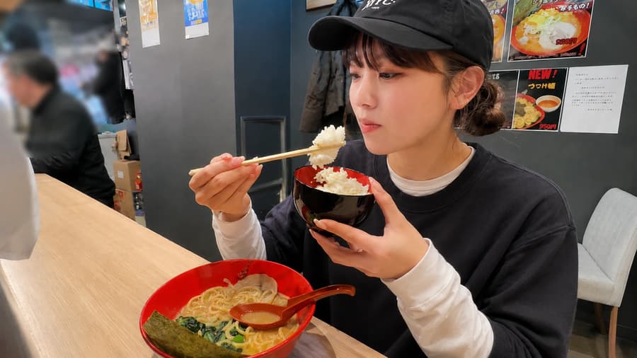 Mika lifting a bite of rice beside a steaming bowl of ramen at a bright counter