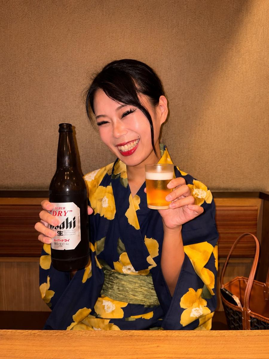 Momoko in a navy and yellow yukata, laughing with a bottle of beer and a glass at an izakaya table