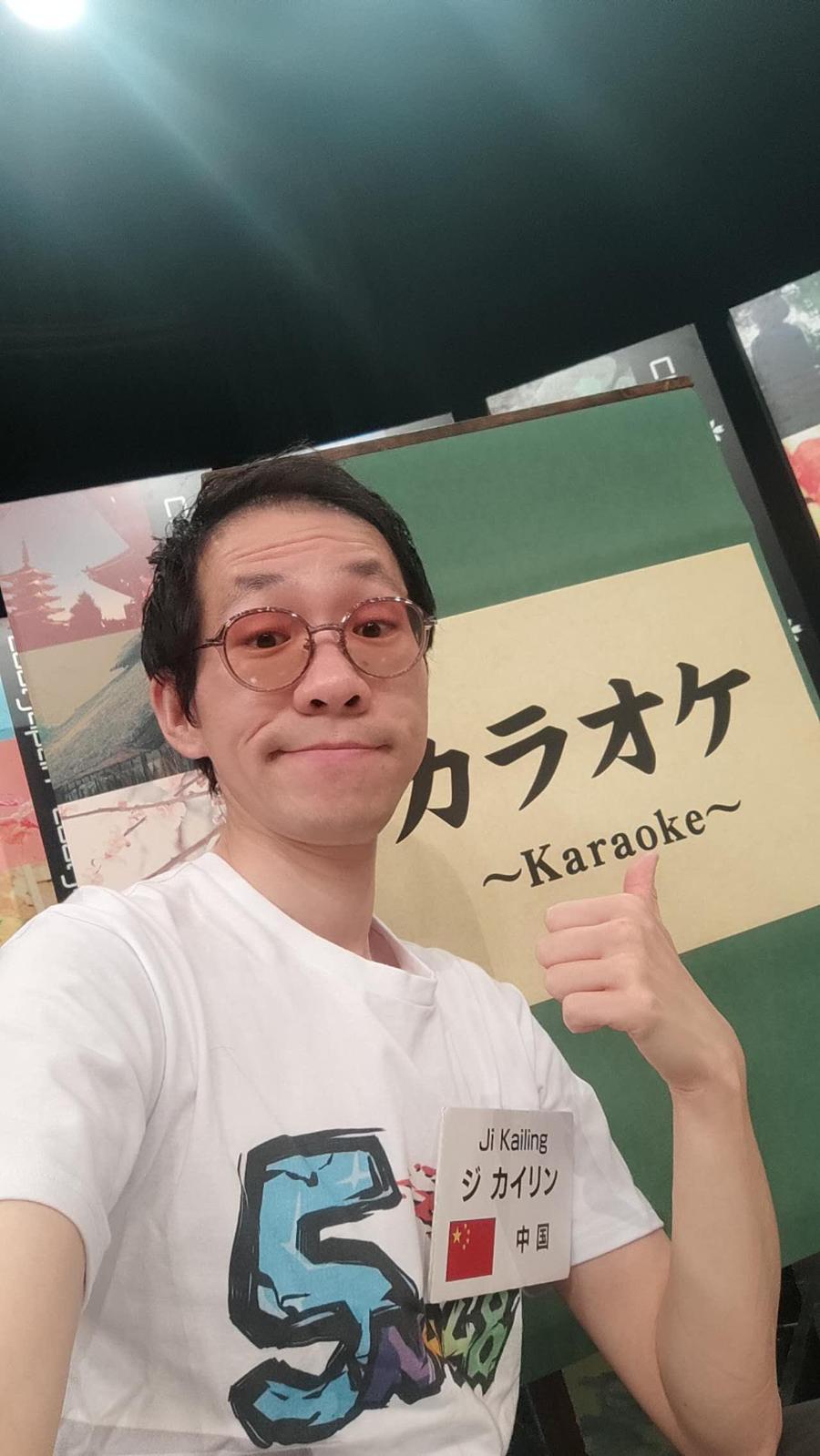 Kailing giving a thumbs up beside a karaoke sign, wearing a name badge