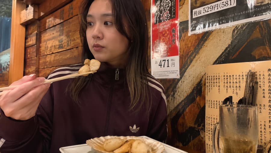 Nanami lifting a piece of food on chopsticks beside a plate served on a scallop shell at an izakaya counter