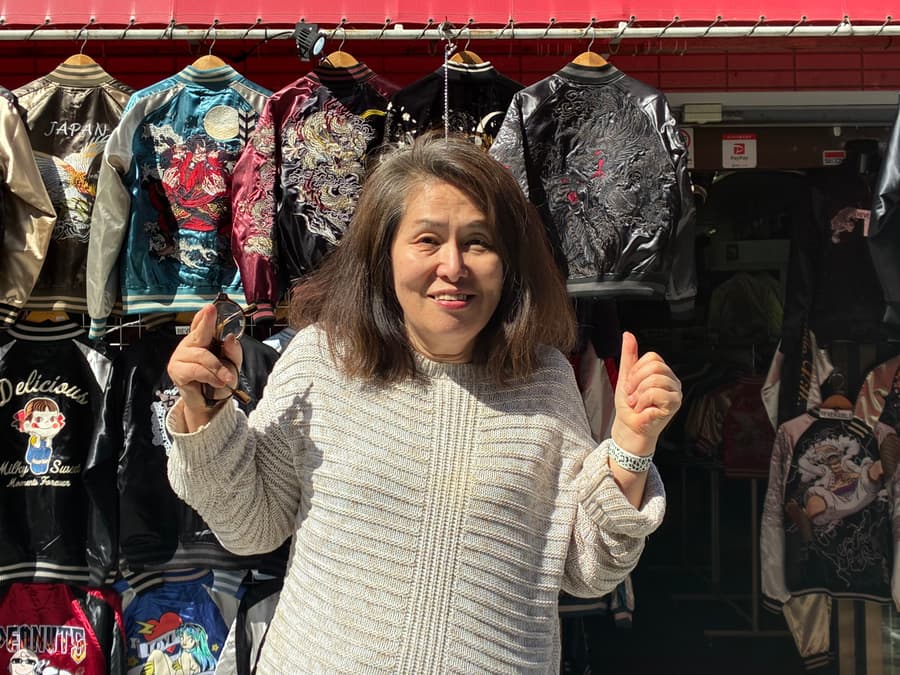 Motoko giving two thumbs up in front of a rack of embroidered sukajan jackets on Dobuita Street