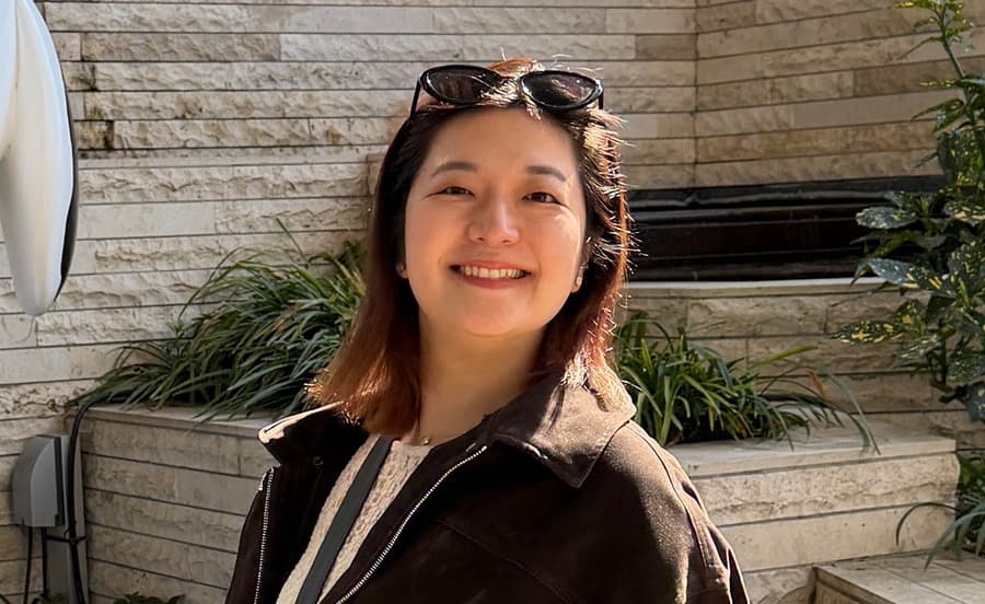 MIYU smiling with sunglasses pushed up on her head, outside a stone-walled building