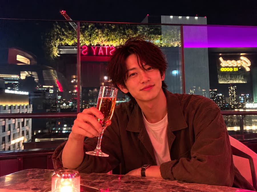 Sora raising a glass of sparkling wine at a rooftop bar with city lights behind him