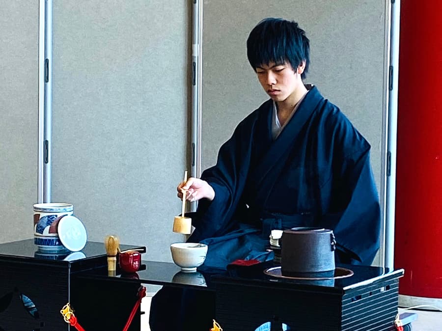 Haruki in a navy kimono ladling hot water into a tea bowl at a tea ceremony
