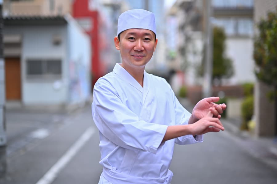 Satoshi in a white sushi chef uniform and cap shaping sushi rice on a quiet Tokyo street